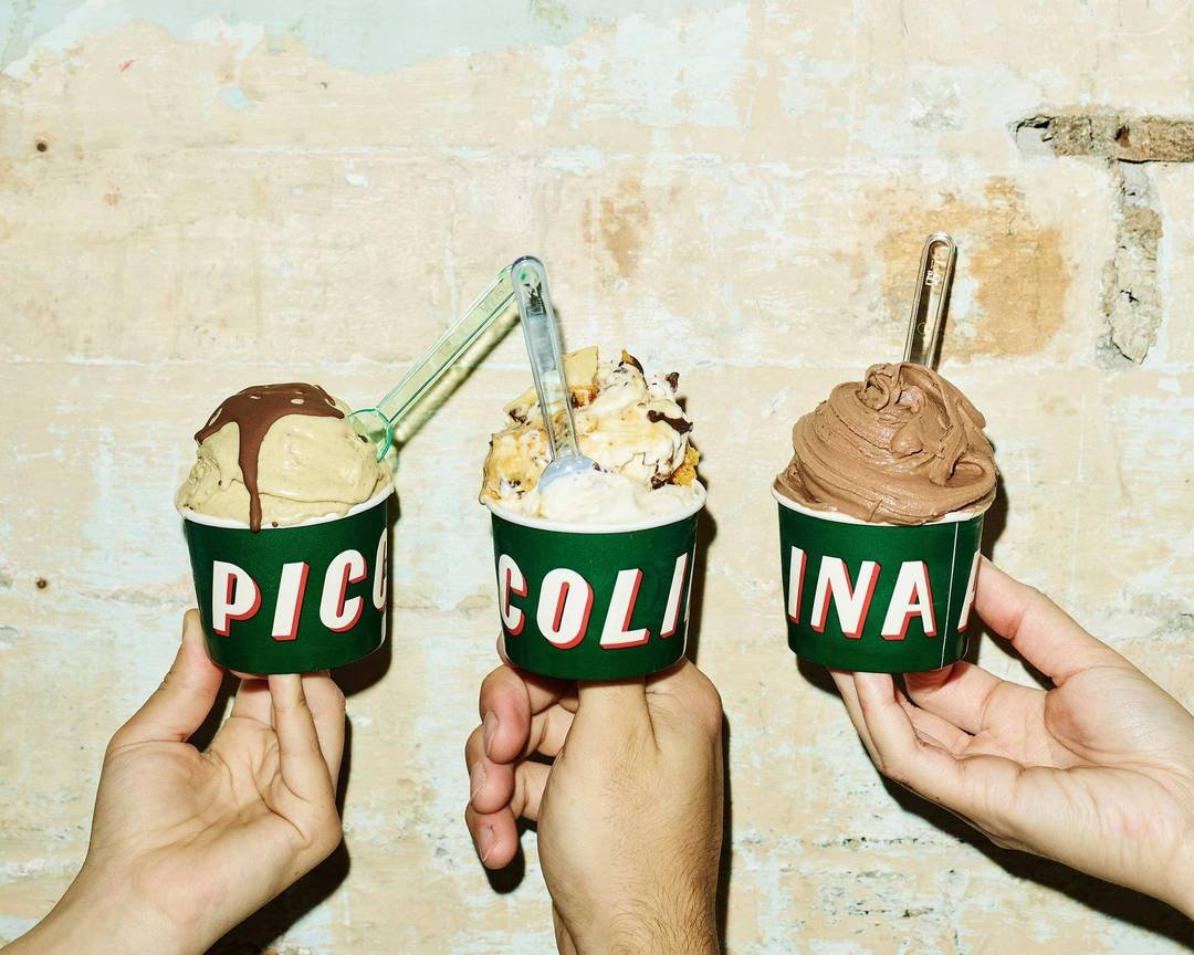 Three hands holding up Piccolina gelato cups against a rustic rendered wall